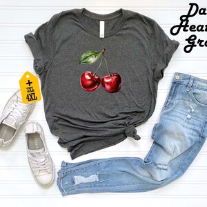 Peut inclure: T-shirt gris chin&eacute; fonc&eacute; avec une illustration &agrave; l'aquarelle de deux cerises rouges et une feuille verte. Le t-shirt est associ&eacute; &agrave; un jean en denim bleu clair et des baskets blanches. L'&eacute;tiquette indique les tailles 3XL et 4XL.