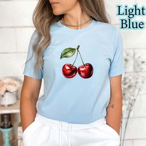 Peut inclure: T-shirt bleu clair avec une illustration &agrave; l'aquarelle de deux cerises rouges et d'une feuille verte. Le texte "Light Blue" est imprim&eacute; en bleu clair en haut &agrave; droite. Le t-shirt est en mati&egrave;re souple.