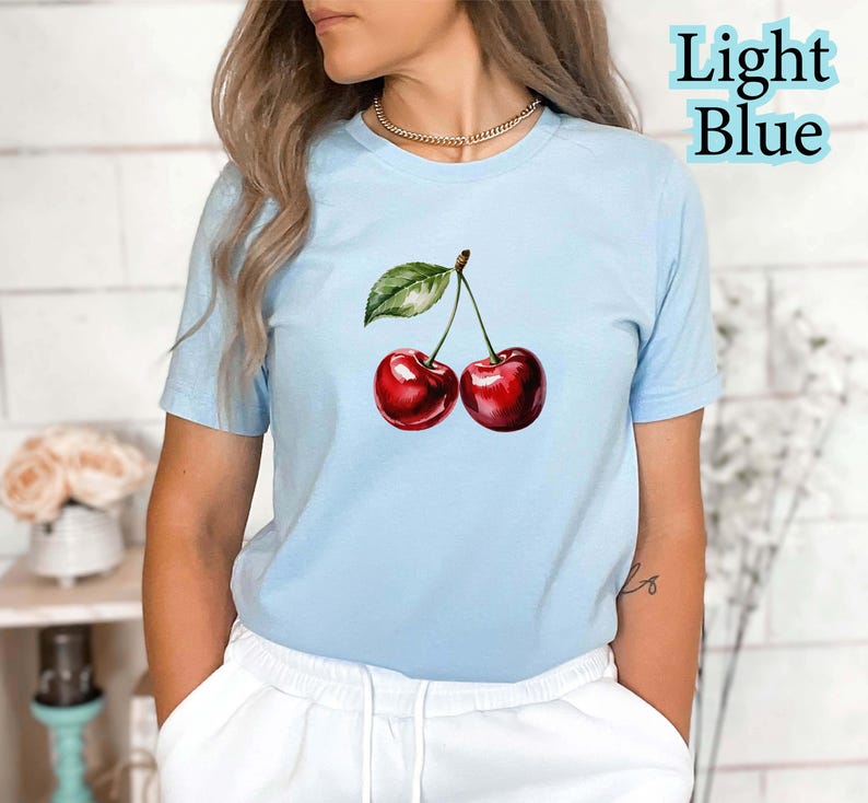 Peut inclure: T-shirt bleu clair avec une illustration &agrave; l'aquarelle de deux cerises rouges et d'une feuille verte. Le texte "Light Blue" est imprim&eacute; en bleu clair en haut &agrave; droite. Le t-shirt est en mati&egrave;re souple.
