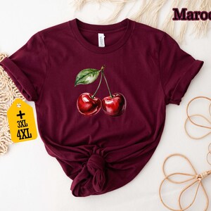 Peut inclure: T-shirt bordeaux avec une illustration &agrave; l'aquarelle de deux cerises et une feuille verte. Le t-shirt a un col rond et des manches courtes. Une &eacute;tiquette avec les tailles 3XL et 4XL est attach&eacute;e.