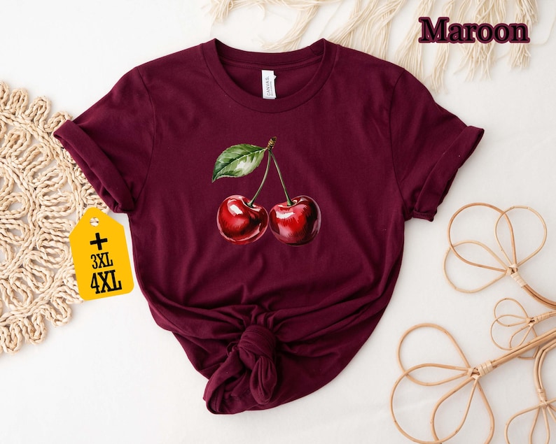 Peut inclure: T-shirt bordeaux avec une illustration &agrave; l'aquarelle de deux cerises et une feuille verte. Le t-shirt a un col rond et des manches courtes. Une &eacute;tiquette avec les tailles 3XL et 4XL est attach&eacute;e.