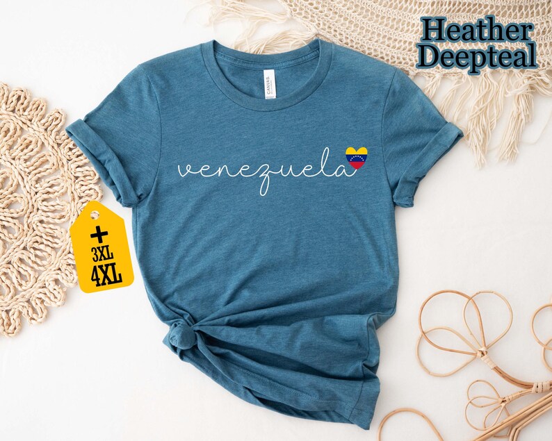 May include: A blue t-shirt with the word "venezuela" in cursive script and a heart-shaped Venezuelan flag graphic. A yellow tag indicates plus sizes 3XL and 4XL. The shirt is made of a soft material.