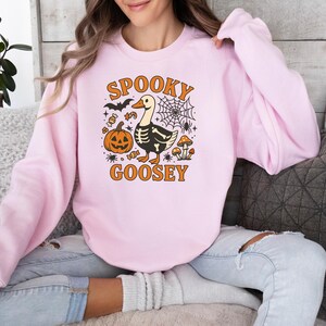 May include: Light pink sweatshirt with a Halloween-themed graphic. The design features a goose with a skeleton body, a jack-o'-lantern, a spiderweb, and the words "Spooky Goosey" in orange. The sweatshirt is a casual, long-sleeved top.