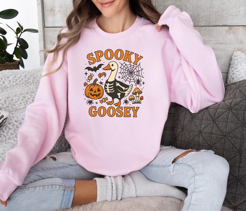 May include: Light pink sweatshirt with a Halloween-themed graphic. The design features a goose with a skeleton body, a jack-o'-lantern, a spiderweb, and the words "Spooky Goosey" in orange. The sweatshirt is a casual, long-sleeved top.