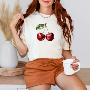 Peut inclure: T-shirt cr&egrave;me avec une illustration &agrave; l'aquarelle de deux cerises m&ucirc;res avec une feuille verte. Les cerises sont d'un rouge profond. Le t-shirt est associ&eacute; &agrave; un short orange. Une tasse blanche est tenue &agrave; la main.