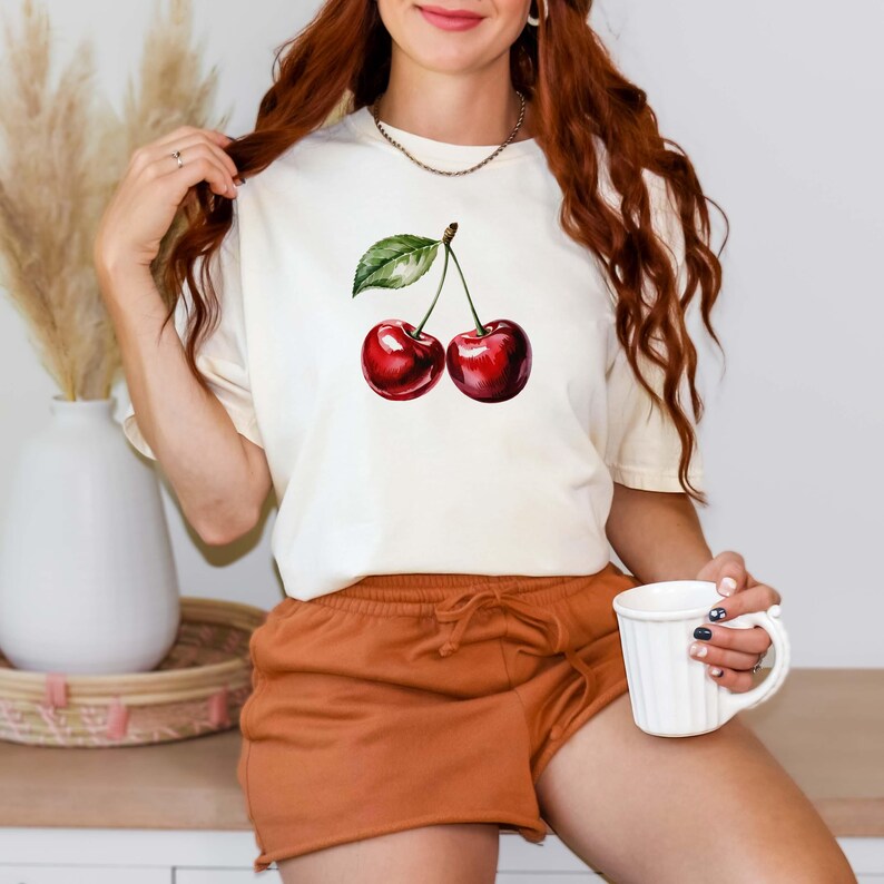 Peut inclure: T-shirt cr&egrave;me avec une illustration &agrave; l'aquarelle de deux cerises m&ucirc;res avec une feuille verte. Les cerises sont d'un rouge profond. Le t-shirt est associ&eacute; &agrave; un short orange. Une tasse blanche est tenue &agrave; la main.