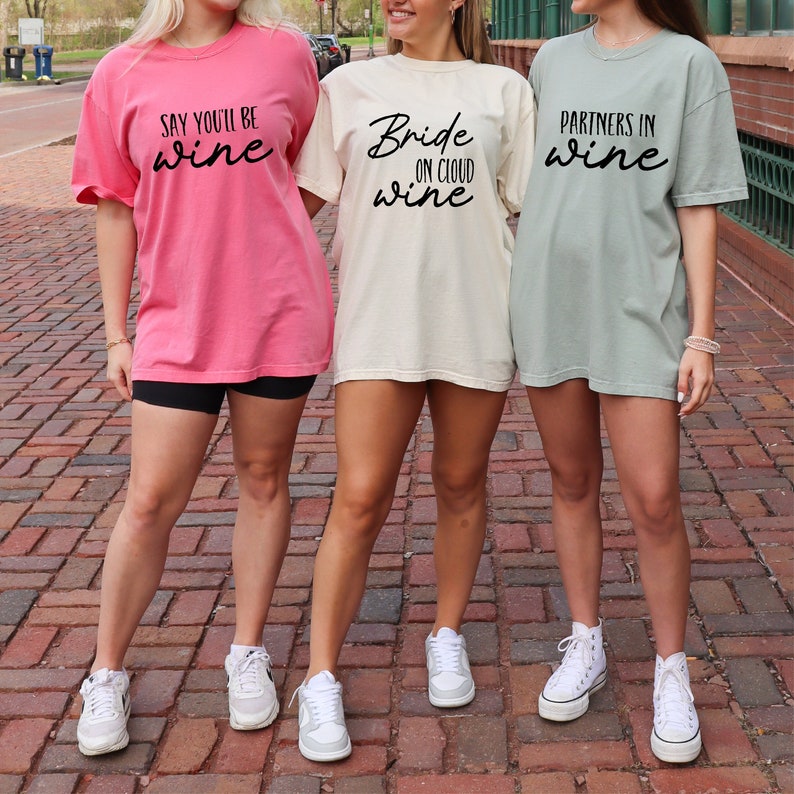 May include: Three women wearing oversized t-shirts with wine-themed phrases. The shirts are in pink, cream, and sage green. The pink shirt says "Say You'll Be Wine", the cream shirt says "Bride on Cloud Wine", and the green shirt says "Partners in Wine".