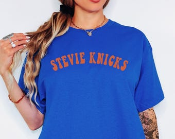 May include: A royal blue short-sleeved t-shirt featuring the text STEVIE KNICKS printed in a bold, orange, arched font across the chest. The garment has a classic crew neckline and a relaxed fit, providing a simple and straightforward design.