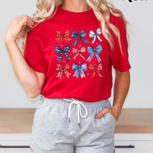 K&ouml;nnte beinhalten: Rotes T-Shirt mit einem Muster aus blauen, wei&szlig;en und roten Schleifen. Die Schleifen sind in einem Raster angeordnet und haben ein Sternendesign. Das Shirt wird von einer Person mit langen blonden Haaren getragen. Die Person tr&auml;gt graue Shorts.