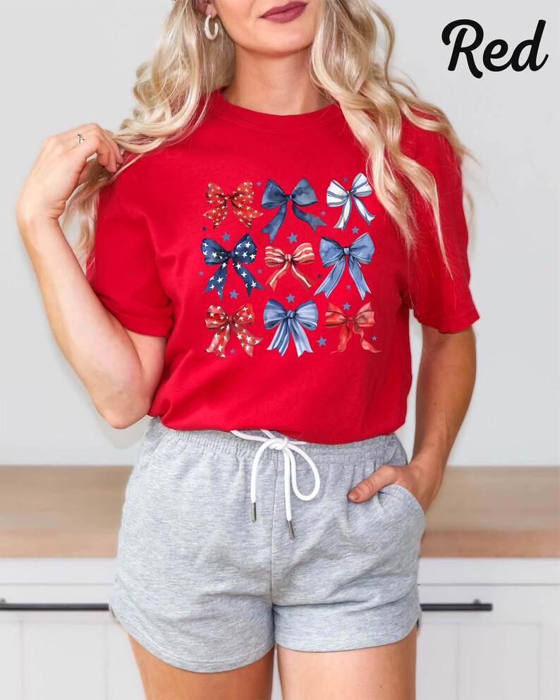 K&ouml;nnte beinhalten: Rotes T-Shirt mit einem Muster aus blauen, wei&szlig;en und roten Schleifen. Die Schleifen sind in einem Raster angeordnet und haben ein Sternendesign. Das Shirt wird von einer Person mit langen blonden Haaren getragen. Die Person tr&auml;gt graue Shorts.