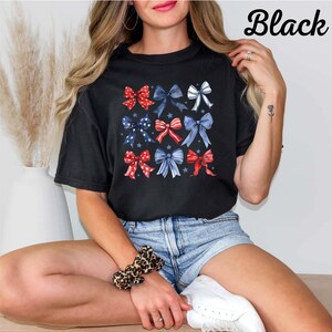 K&ouml;nnte beinhalten: Ein schwarzes T-Shirt mit einem Aufdruck von roten, wei&szlig;en und blauen Schleifen, die in einem Raster angeordnet sind. Die Schleifen sind mit Sternen und Streifen verziert.