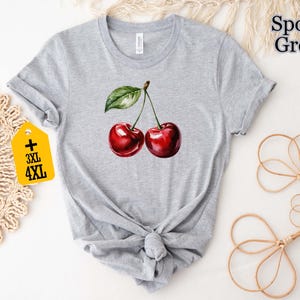 Peut inclure: T-shirt gris clair avec une illustration &agrave; l'aquarelle de deux cerises m&ucirc;res. Les cerises sont d'un rouge profond avec une feuille et une tige vertes. Le t-shirt porte le texte "Sport Grey" et une &eacute;tiquette "3XL 4XL".