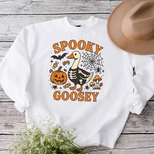 May include: White sweatshirt with a Halloween-themed graphic. The design features a goose with a skeleton outline, a pumpkin, bats, candy, and the words "Spooky Goosey" in orange. A brown hat is visible in the background.