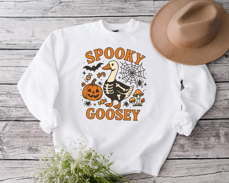 May include: White sweatshirt with a Halloween-themed graphic. The design features a goose with a skeleton outline, a pumpkin, bats, candy, and the words "Spooky Goosey" in orange. A brown hat is visible in the background.