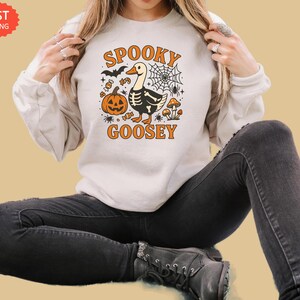 May include: Cream-colored sweatshirt with a Halloween-themed graphic. The design features a cartoon goose with a skeleton body, a jack-o'-lantern, a spider web, and the words "Spooky Goosey" in orange.
