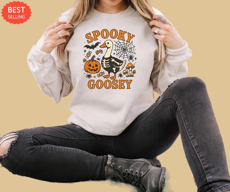 May include: Cream-colored sweatshirt with a Halloween-themed graphic. The design features a cartoon goose with a skeleton body, a jack-o'-lantern, a spider web, and the words "Spooky Goosey" in orange.