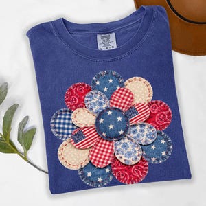 May include: A blue Comfort Colors t-shirt featuring a large flower design. The flower is made of various fabric patterns, including the American flag, gingham, and floral prints. The flower's center has white stars on a blue background.