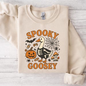 May include: Beige sweatshirt with a Halloween-themed graphic. The design features a goose with a skeleton body, a jack-o'-lantern, a spider web, and the words "Spooky Goosey" in orange. Additional elements include bats, candy, spiders, and mushrooms.