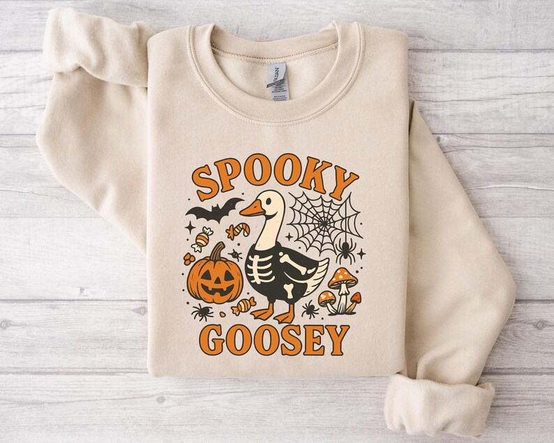 May include: Beige sweatshirt with a Halloween-themed graphic. The design features a goose with a skeleton body, a jack-o'-lantern, a spider web, and the words "Spooky Goosey" in orange. Additional elements include bats, candy, spiders, and mushrooms.