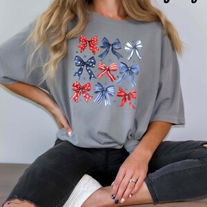 K&ouml;nnte beinhalten: Ein graues T-Shirt mit einem Design aus roten, wei&szlig;en und blauen Schleifen. Die Schleifen sind in einem Raster angeordnet und haben einen Aquarell-Effekt. Das Wort "Grey" ist in wei&szlig;er Schrift in der oberen rechten Ecke des Bildes geschrieben.