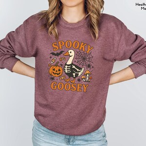 May include: Maroon sweatshirt with a Halloween-themed graphic. The design features a goose with a skeleton body, a jack-o'-lantern, spiderwebs, and the words "Spooky Goosey" in orange. A bat and candy corn complete the design.