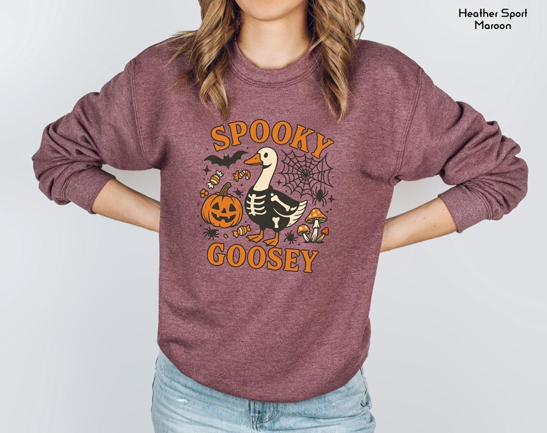 May include: Maroon sweatshirt with a Halloween-themed graphic. The design features a goose with a skeleton body, a jack-o'-lantern, spiderwebs, and the words "Spooky Goosey" in orange. A bat and candy corn complete the design.