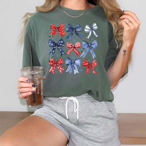 K&ouml;nnte beinhalten: Ein gr&uuml;nes T-Shirt mit kurzen &Auml;rmeln und einem Muster aus roten, wei&szlig;en und blauen Schleifen. Die Schleifen sind in einem Raster angeordnet und haben einen Aquarell-Effekt. Das Shirt wird mit grauen Shorts kombiniert.