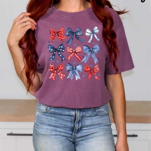 K&ouml;nnte beinhalten: Ein lilafarbenes T-Shirt mit einem Muster aus roten, wei&szlig;en und blauen Schleifen. Die Schleifen sind in einem Raster angeordnet und haben einen Aquarell-Effekt. Der Text "Berry" befindet sich in der oberen rechten Ecke des Bildes.