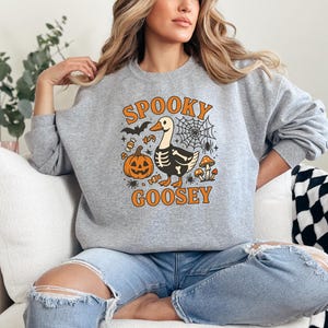 May include: A gray sweatshirt with a Halloween-themed graphic. The design features a cartoon goose with a skeleton body, a jack-o'-lantern, spiderwebs, and the words "Spooky Goosey" in orange. The sweatshirt is long-sleeved.