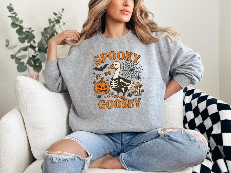 May include: A gray sweatshirt with a Halloween-themed graphic. The design features a cartoon goose with a skeleton body, a jack-o'-lantern, spiderwebs, and the words "Spooky Goosey" in orange. The sweatshirt is long-sleeved.