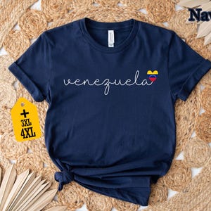 May include: Navy blue t-shirt with the word "Venezuela" in white script and a heart-shaped flag graphic. A yellow tag indicates plus sizes 3XL and 4XL. The shirt is displayed on a woven, neutral-toned surface.