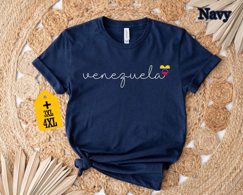 May include: Navy blue t-shirt with the word "Venezuela" in white script and a heart-shaped flag graphic. A yellow tag indicates plus sizes 3XL and 4XL. The shirt is displayed on a woven, neutral-toned surface.