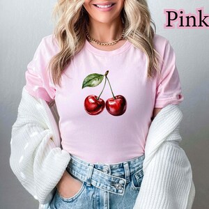 Peut inclure: T-shirt rose clair avec une illustration &agrave; l'aquarelle de deux cerises et une feuille verte. Le mot "Pink" est imprim&eacute; en rose en haut &agrave; droite. La personne porte un jean bleu et un pull blanc.