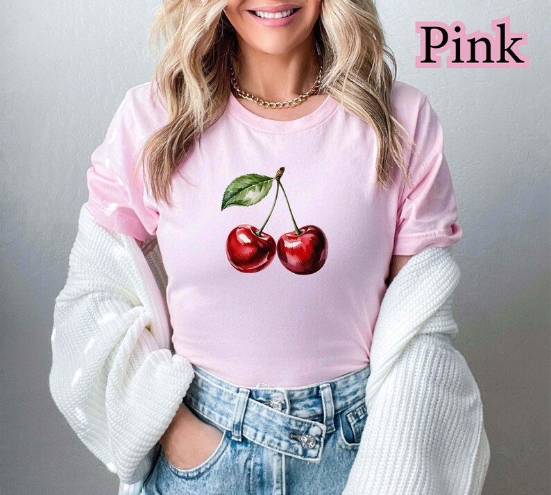 Peut inclure: T-shirt rose clair avec une illustration &agrave; l'aquarelle de deux cerises et une feuille verte. Le mot "Pink" est imprim&eacute; en rose en haut &agrave; droite. La personne porte un jean bleu et un pull blanc.