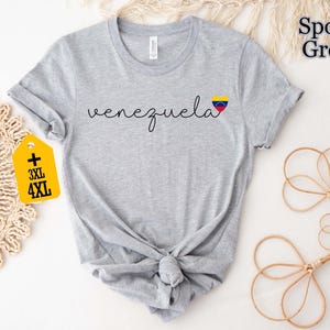 May include: A light gray t-shirt with the word "venezuela" in a cursive font and a heart-shaped flag graphic. The shirt has a knotted hem and a tag indicating 3XL and 4XL sizes. The words "Sport Grey" are printed on the top right.