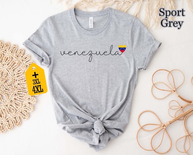 May include: A light gray t-shirt with the word "venezuela" in a cursive font and a heart-shaped flag graphic. The shirt has a knotted hem and a tag indicating 3XL and 4XL sizes. The words "Sport Grey" are printed on the top right.