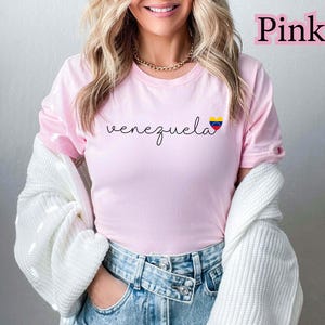 May include: A light pink t-shirt with the word "venezuela" in a cursive font. The shirt features a small heart-shaped graphic with the Venezuelan flag colors. The word "Pink" is written in a pink font in the upper right corner.