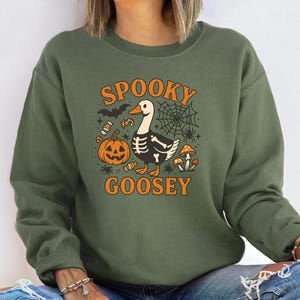 May include: Olive green sweatshirt with the text "SPOOKY GOOSEY" in orange. The graphic features a goose with a skeleton design, a jack-o'-lantern, spiderwebs, bats, candy, and mushrooms. A fun Halloween-themed design.