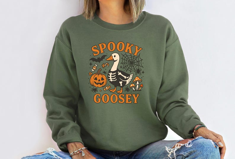 May include: Olive green sweatshirt with the text "SPOOKY GOOSEY" in orange. The graphic features a goose with a skeleton design, a jack-o'-lantern, spiderwebs, bats, candy, and mushrooms. A fun Halloween-themed design.