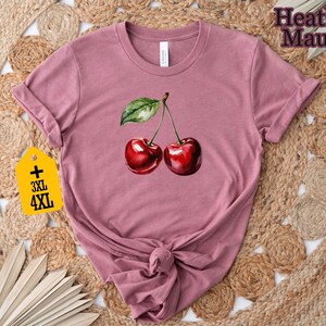 Peut inclure: T-shirt mauve chin&eacute; avec une illustration &agrave; l'aquarelle de deux cerises m&ucirc;res et une feuille verte. Les manches sont retrouss&eacute;es et l'ourlet est nou&eacute;. Une &eacute;tiquette jaune indique les grandes tailles 3XL et 4XL.