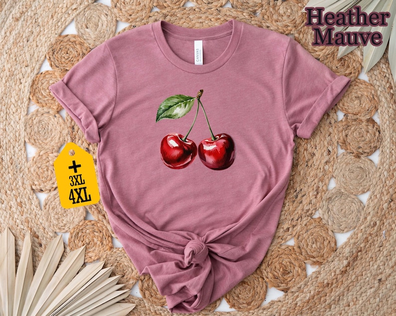 Peut inclure: T-shirt mauve chin&eacute; avec une illustration &agrave; l'aquarelle de deux cerises m&ucirc;res et une feuille verte. Les manches sont retrouss&eacute;es et l'ourlet est nou&eacute;. Une &eacute;tiquette jaune indique les grandes tailles 3XL et 4XL.