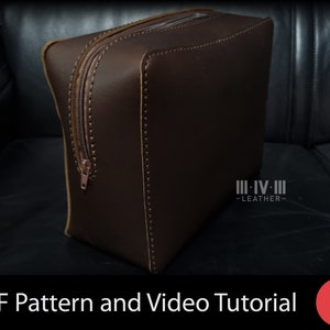May include: A brown leather zippered bag with a tan leather trim. The bag is sitting on a black leather surface. The bag has the text "III-IV-III -LEATHER- " on the side.