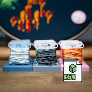 Puede incluir: Tres carretes de hilo de bordar en blanco, gris y naranja, cada uno etiquetado con un número. Los carretes se asientan sobre bases individuales de colores. También hay un icono de archivo digital con las letras "STL".
