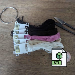 May include: A collection of embroidery floss in black, pink, cream, and white, with a silver metal ring and small scissors. A digital file icon with the letters "STL" is also present. The items are arranged on a wooden surface.