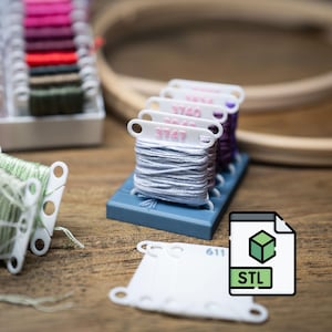 May include: A collection of embroidery floss in various colors, including gray, purple, and green, displayed on white and blue plastic organizers. An embroidery hoop and a digital file icon with "STL" text are also visible.