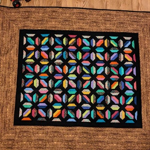 May include: A colorful quilt with a black center featuring a geometric pattern of various colored shapes. The quilt has a brown border with a wood grain design, and a black border. The quilt is on a wooden floor.