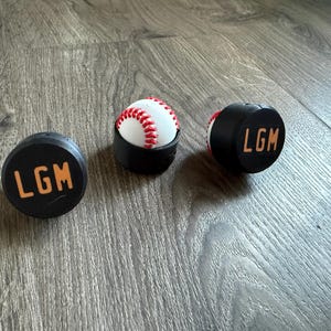 May include: Three baseball-themed items on a wooden surface. A baseball sits in a black holder, flanked by two black puck-shaped objects with "LGM" in orange lettering. The baseball has red stitching.
