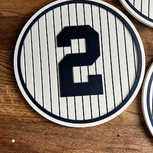 May include: A white and black baseball-themed wall decor with a black number "2" in the center. The decor is round and has a white background with black stripes.