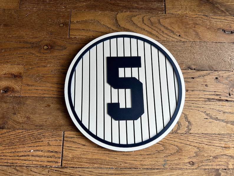 May include: A white and blue baseball-themed sign with a black number 5 in the center. The sign has a white background with blue stripes and a blue border.