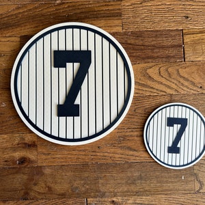 May include: Two round wooden signs with a baseball field design and the number "7" in black. The larger sign is approximately 12 inches in diameter and the smaller sign is approximately 6 inches in diameter.